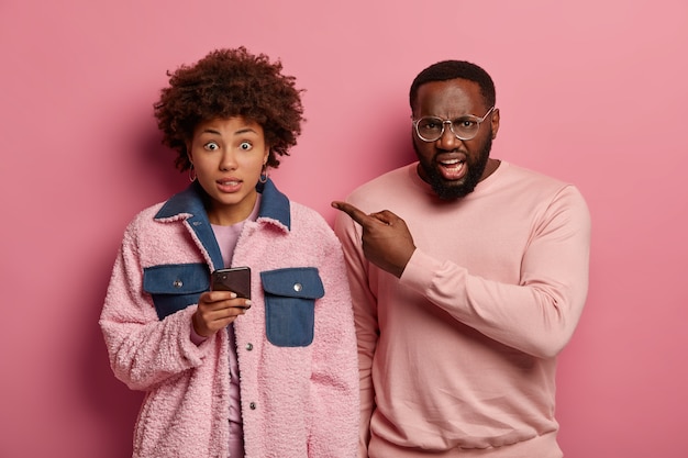 Un homme mécontent en colère à lunettes montre une femme afro-américaine avec un smartphone qui a l'air coupable et dit oups, accro aux technologies modernes. Couple ethnique pose ensemble à l'intérieur, étant très émotif