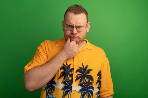 Homme mécontent à lunettes et chemise orange fronçant les sourcils debout sur mur vert