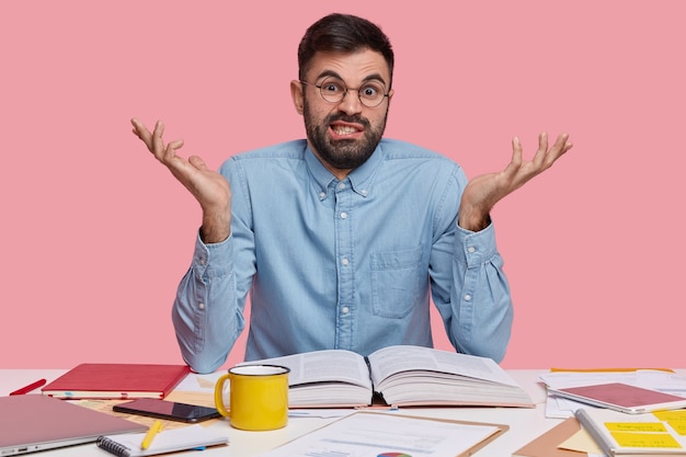 Photo gratuite un homme mécontent mal rasé en colère écarte les mains, se sent insatisfait et désemparé, lit l'encyclopédie, s'assoit à l'espace de travail, boit du thé