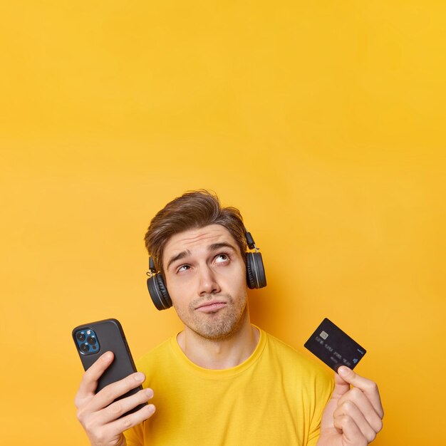 Un homme mécontent réfléchi concentré au-dessus écoute de la musique via un casque tient un téléphone portable et une carte de crédit pense comment effectuer un paiement sur un site Web isolé sur un espace vide de mur jaune au-dessus