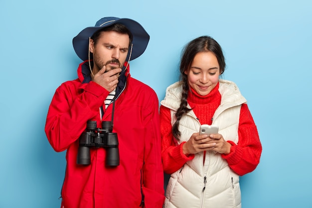 Un homme mécontent tient le menton, regarde avec colère le smartphone de sa petite amie, vêtu de vêtements décontractés, porte des jumelles et un message de type fille asiatique heureuse, concentré sur cellulaire