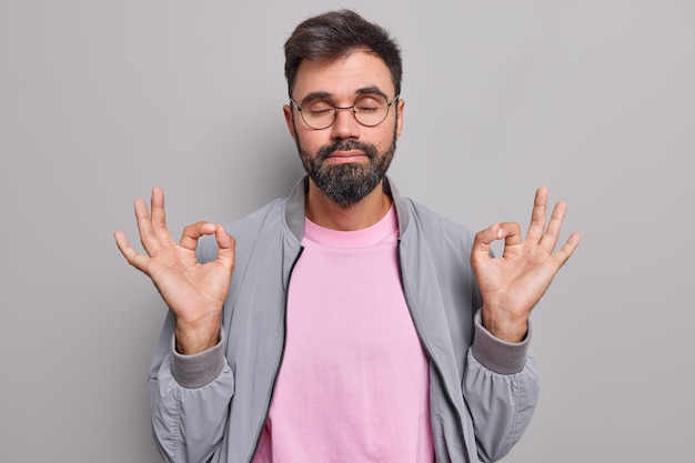 l'homme médite seul fait un geste correct avec les deux mains respire profondément garde les yeux fermés pratique le yoga porte des lunettes rondes veste grise t-shirt rose