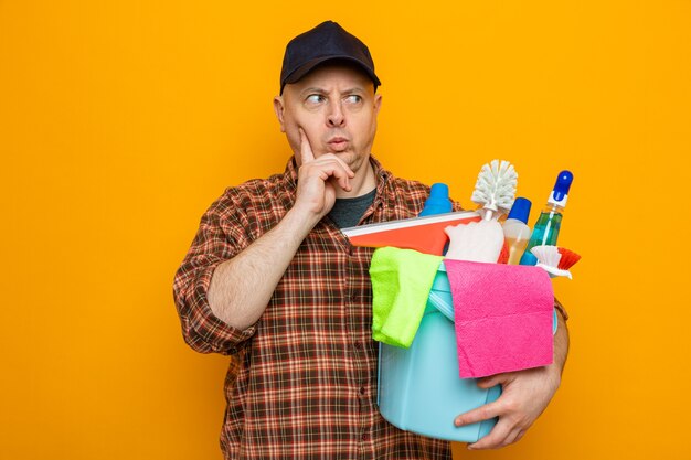 Homme de ménage en chemise à carreaux et casquette tenant un seau avec des outils de nettoyage à côté avec un visage renfrogné perplexe debout sur fond orange