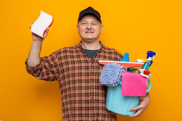 Homme de ménage en chemise à carreaux et casquette tenant un seau avec des outils de nettoyage et une éponge regardant avec le sourire sur le visage