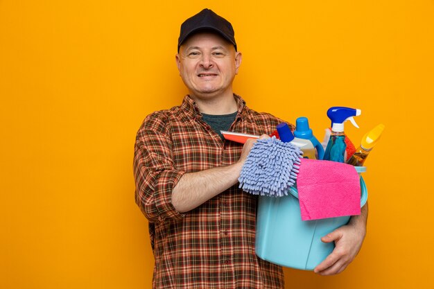 Homme de ménage en chemise à carreaux et casquette tenant un seau avec des outils de nettoyage regardant avec le sourire sur le visage heureux et positif