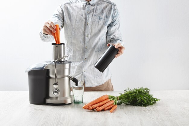 L'homme met les carottes à l'intérieur d'un presse-agrumes métallique avec un verre vide pour faire un jus savoureux pour le petit déjeuner