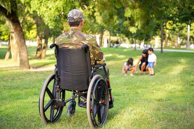 Homme militaire à la retraite handicapé solitaire en fauteuil roulant en regardant sa femme et ses petits enfants jouant ensemble dans le parc. Vue arrière. Concept de vétéran de guerre ou d'invalidité