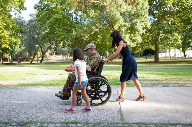 Homme militaire souriant marchant avec la famille dans le parc de la ville. Mère aux cheveux longs poussant un fauteuil roulant. Petite fille qui marche et parle avec papa handicapé. Concept de famille en plein air, week-end et handicap