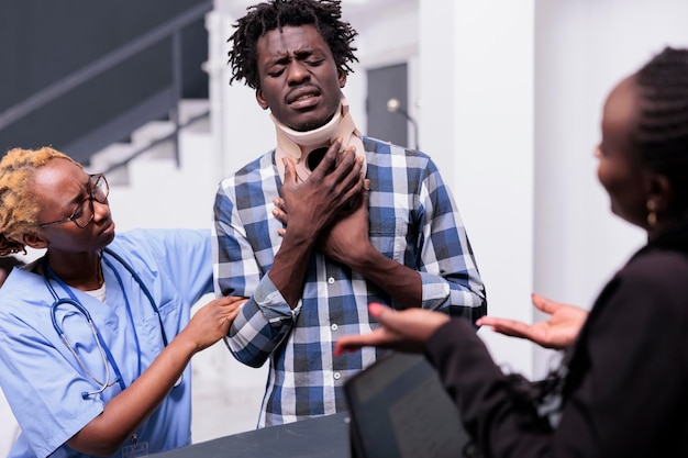 Homme avec une minerve médicale recevant le soutien et l'aide d'une infirmière et d'une réceptionniste, donnant un traitement pour soigner la douleur après une blessure accidentelle. Personne portant de la mousse de collier cervical à la réception de l'hôpital.