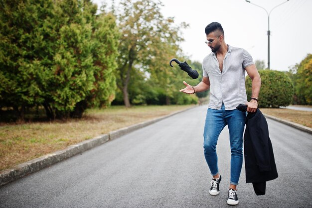 Homme à la mode grand barbe arabe porter sur chemise jeans et lunettes de soleil marchant au parc avec parapluie et manteau à portée de main