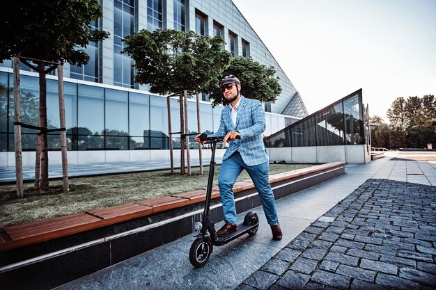 Un homme moderne et joyeux conduit son scooter électrique près de son bureau après une longue journée difficile.