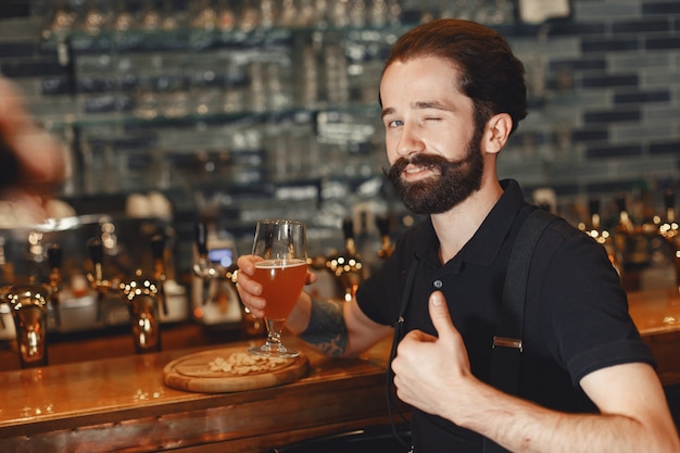 Un homme avec une moustache et une barbe se tient au bar et boit de l'alcool dans un verre.