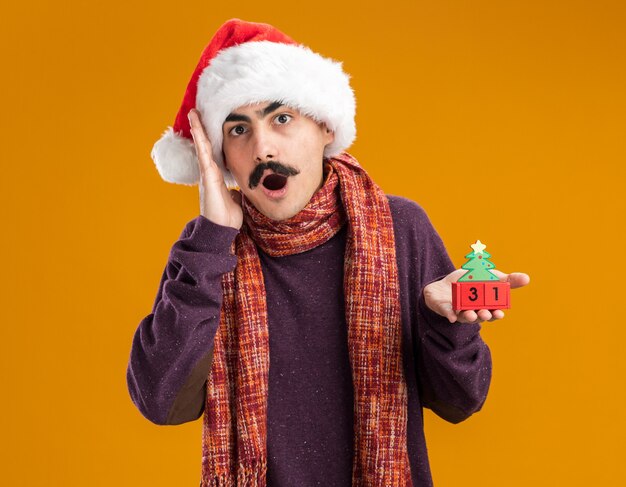 Homme moustachu portant un bonnet de noel avec une écharpe chaude autour du cou tenant des cubes de jouets avec la date du nouvel an étonné et surpris debout sur un mur orange