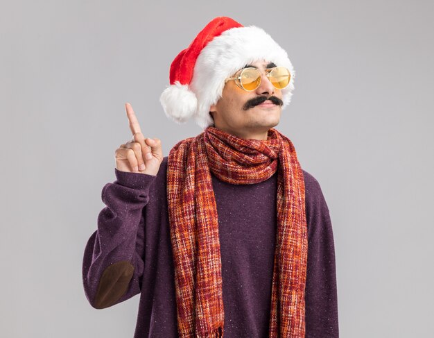 Homme moustachu portant un bonnet de noel et des lunettes jaunes avec une écharpe chaude autour du cou levant les yeux avec le sourire sur le visage montrant l'index debout sur un mur blanc