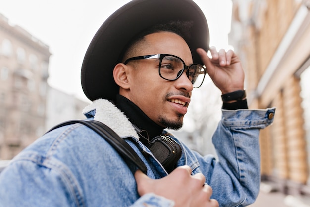 L'homme mulâtre inspiré porte une veste en jean décontractée marchant dans la rue. Photo extérieure d'un homme africain au chapeau noir et lunettes élégantes, passer du temps dans la ville.