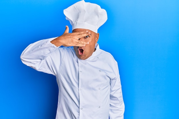 Photo gratuite homme mûr du moyen-orient portant un uniforme de cuisinier professionnel et un chapeau regardant sous le choc couvrant le visage et les yeux avec la main, regardant à travers les doigts avec une expression embarrassée.