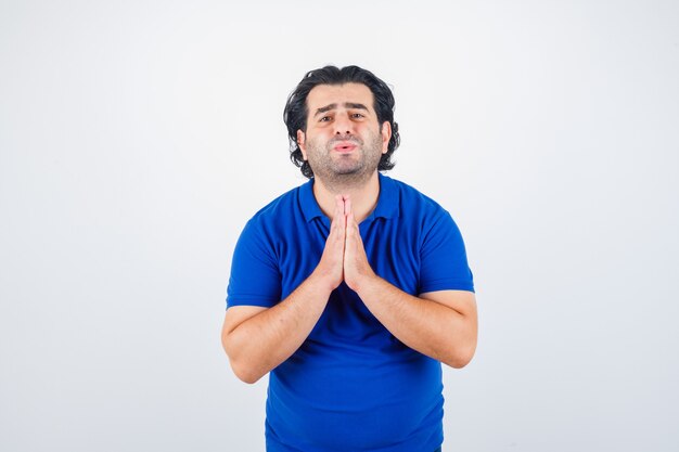 Homme mûr en t-shirt bleu, serrant les mains en position de prière et à la déçu, vue de face.