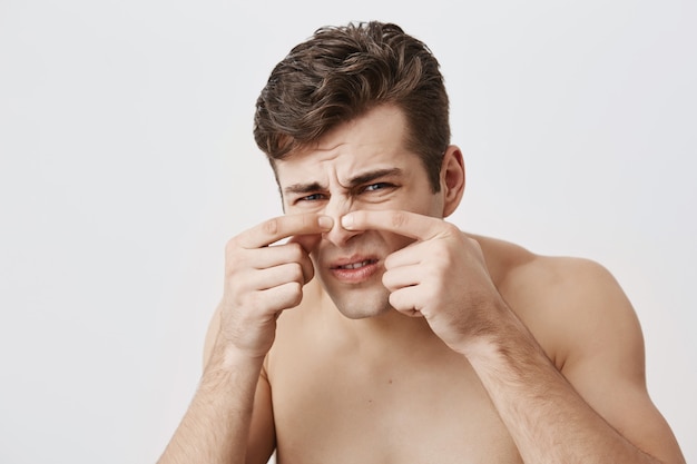 Homme musclé nu avec une apparence attrayante, visage froncé, nez touchant, ayant des problèmes de peau. Homme de race blanche aux cheveux noirs, regardant avec ses yeux bleus. Concept de beauté et de jeunesse.