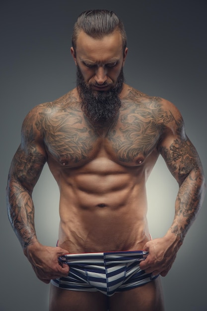 Homme musclé tatoué avec barbe en culotte à rayures posant dans les ombres sur fond gris.