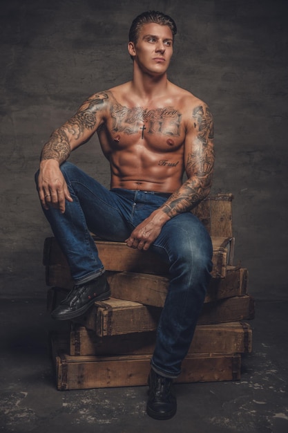 Homme musclé torse nu avec des tatouages sur son corps assis sur des caisses en bois.