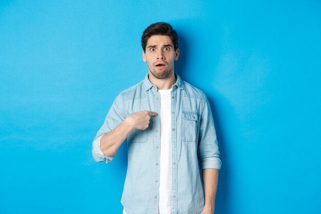 Homme nerveux pointant sur lui-même et ayant l'air confus, debout dans des vêtements décontractés sur fond bleu.