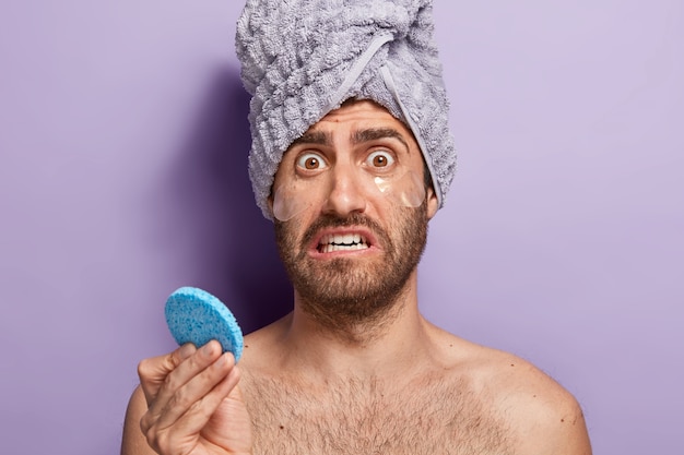 Un homme nerveux regarde avec une expression de visage mécontente inquiète, tient une éponge cosmétique, applique des patchs d'hydrogel pour enlever les poches sous les yeux