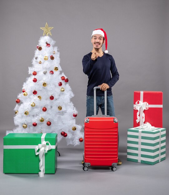 Homme de Noël avec bonnet de noel tenant une valise rouge près de l'arbre de Noël blanc avec des jouets de Noël colorés