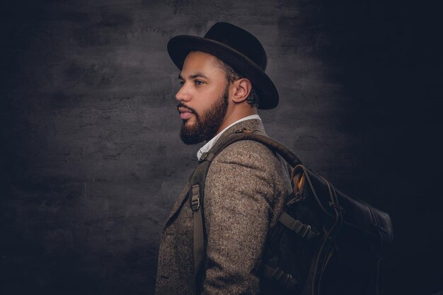 Homme noir barbu vêtu d'un costume en laine et d'un chapeau de feutre tenant un sac à dos.