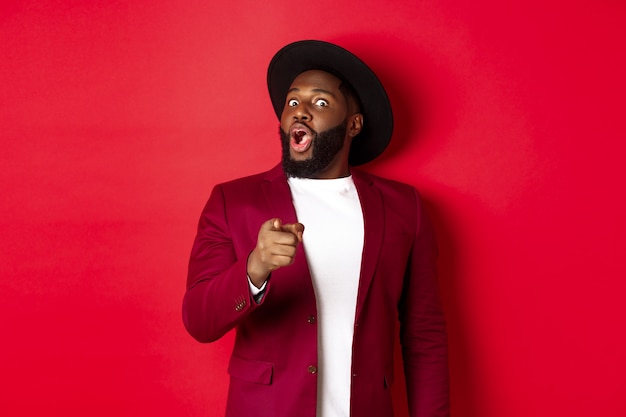 Un homme noir choqué haletant étonné et pointant le doigt vers la caméra, reconnaît quelqu'un, debout dans un blazer rouge et un chapeau sur fond de studio.