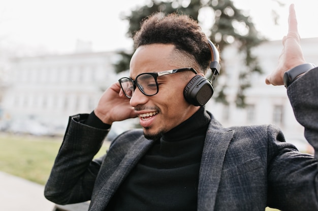 Homme noir avec une coiffure africaine à la mode, écouter de la musique sur la ville. Beau mec mulâtre en veste décontractée et casque posant dans le parc.