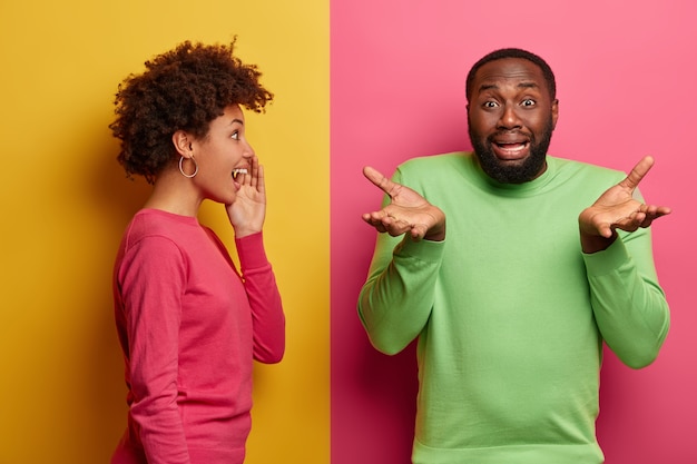 Photo gratuite un homme noir déçu écarte les paumes, a l'air hésitant et malheureux, fait face à une situation problématique, une femme afro-américaine positive murmure un secret à son petit ami, se tient sur le côté. couleur rose et jaune