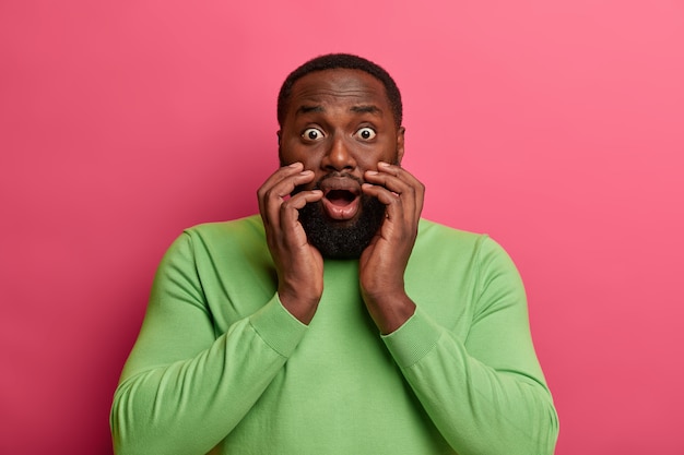 Un homme noir effrayé et effrayé regarde avec des yeux ouverts, assiste à une catastrophe, halète de choc, ouvre largement la bouche, porte un pull vert