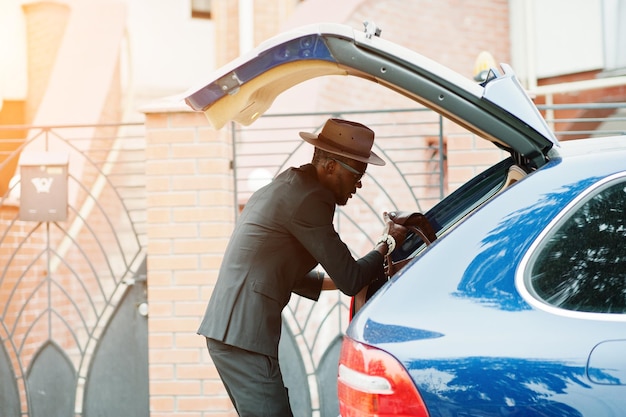 Homme noir élégant à lunettes avec chapeau porter sur costume avec sac à main contre voiture de luxe Riche homme d'affaires afro-américain