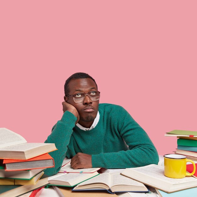 Un homme noir ennuyé réfléchi tient la main sur la joue, regarde vers le haut, porte un pull vert, des lunettes optiques, pense à créer un nouveau projet