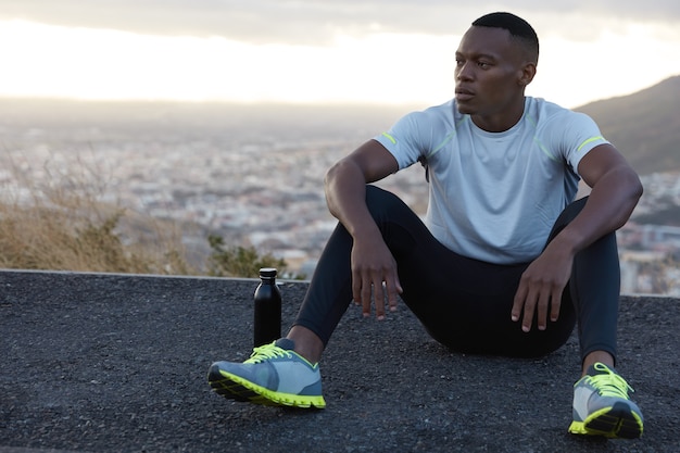 Un homme noir pensif insouciant s'appuie les mains sur les genoux, repose sur l'asphalte, boit de l'eau fraîche, respire l'air, aime la solitude, modèles sur vue floue panoramique avec espace de copie pour votre publicité