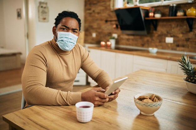 Homme noir portant un masque facial tout en utilisant une tablette numérique à la maison et en regardant la caméra