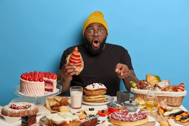 Homme noir stupéfait mange un délicieux croissant, pointe à table pleine de délicieux desserts sucrés, porte un chapeau et un t-shirt, pose sur fond bleu