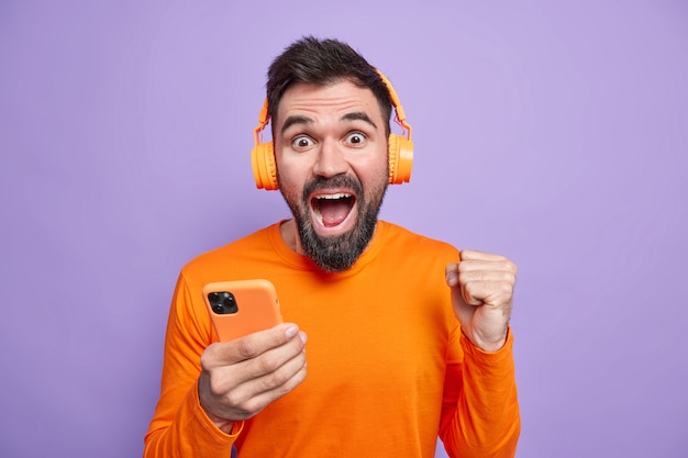 Un Homme Non Rasé Et Ravi Célèbre D'excellentes Nouvelles Serre Le Poing Tient Un Téléphone Portable Utilise Des écouteurs Sans Fil Pour écouter De La Musique Aime Un Bon Son Porte Un Pull Orange