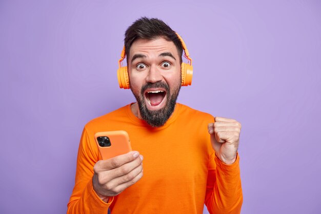 Un homme non rasé et ravi célèbre d'excellentes nouvelles serre le poing tient un téléphone portable utilise des écouteurs sans fil pour écouter de la musique aime un bon son porte un pull orange