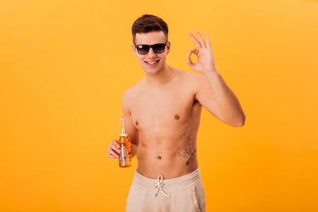 Homme nu souriant en short et lunettes de soleil tenant une bouteille de bière et montrant un signe ok sur jaune