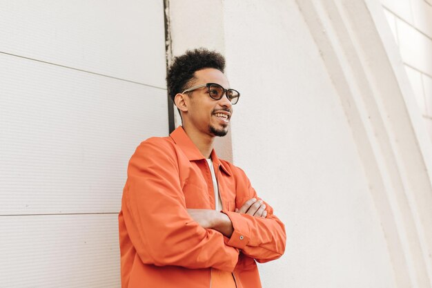 Homme à la peau foncée barbu bouclé de bonne humeur dans une élégante veste orange et des lunettes de soleil sourit croise les bras et s'appuie sur un mur blanc à l'extérieur