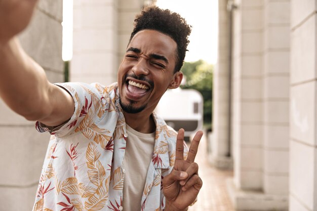 L'homme à la peau foncée bouclé brun fou émotionnel en chemise à fleurs blanche chante montre le signe de la paix et prend le selfie à l'extérieur