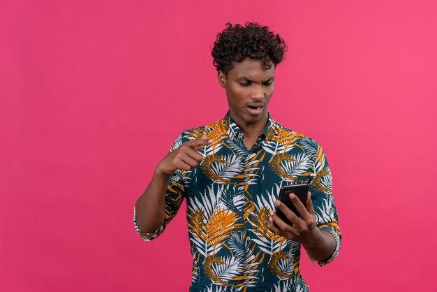 Homme à la peau sombre en colère et nerveux en chemise imprimée de feuilles à la recherche de téléphone mobile et pointant avec l'index