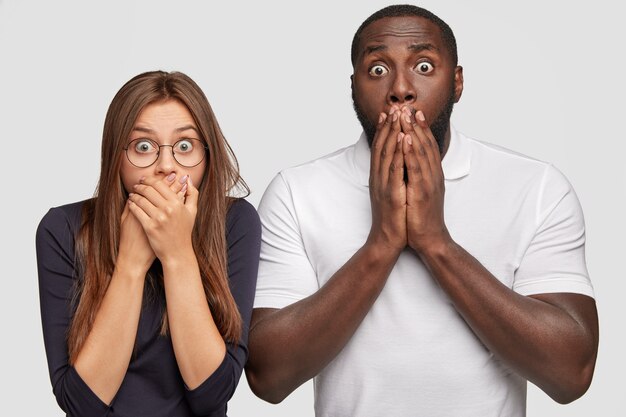 Un homme à la peau sombre et une femme européenne anxieux se couvrent la bouche avec des paumes