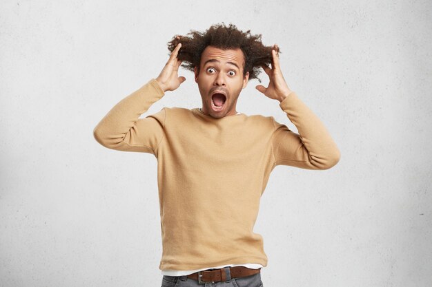 Homme à la peau sombre et terrifié pétrifié avec les cheveux surélevés, regarde avec les yeux sautés et la bouche grande ouverte