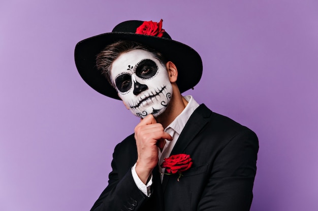 Homme pensif au chapeau noir élégant posant avec un maquillage de fête effrayant. Photo de Studio de beau garçon zombie isolé sur fond clair.