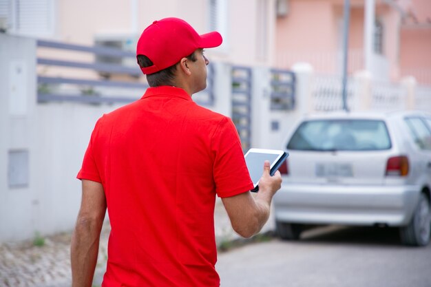 Homme pensif tenant la tablette, debout et à la recherche d'adresse. Courrier professionnel caucasien en uniforme rouge marchant dans la rue.