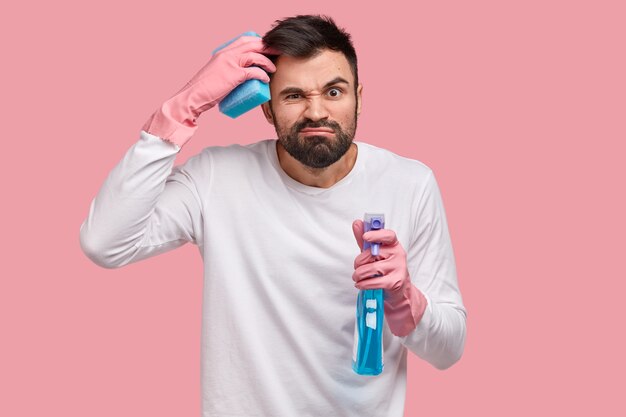 Un homme perplexe se gratte la tête, ne peut pas décider quoi nettoyer en premier, tient une bouteille de spray, prend soin de l'hygiène, porte une éponge