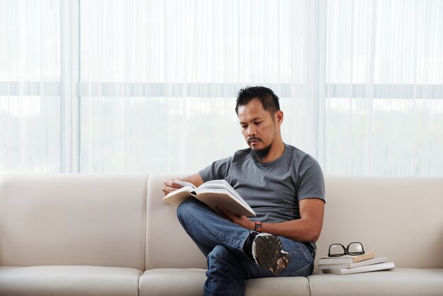 Homme philippin assis sur un canapé avec un pied sur le genou et un livre de lecture