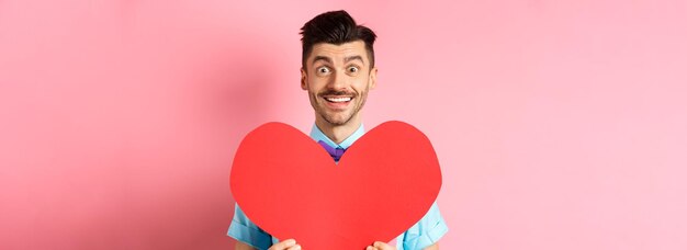 Homme plein d'espoir amoureux montrant un signe de coeur rouge souriant à la caméra attendant l'âme sœur le jour de la saint-valentin